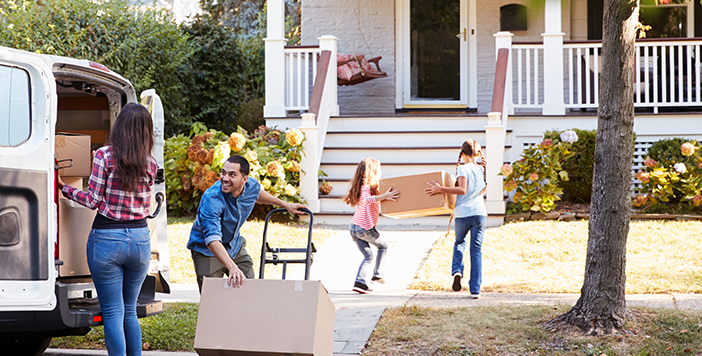 A family moving into a house. Two kids carry a box toward the house, a woman is about to remove a box from the back of a van filled with boxes and a man tilts a dolly with a box on it