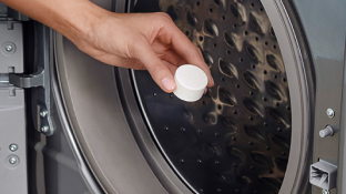 A hand holds an affresh tablet at the opening to the washing machine's drum