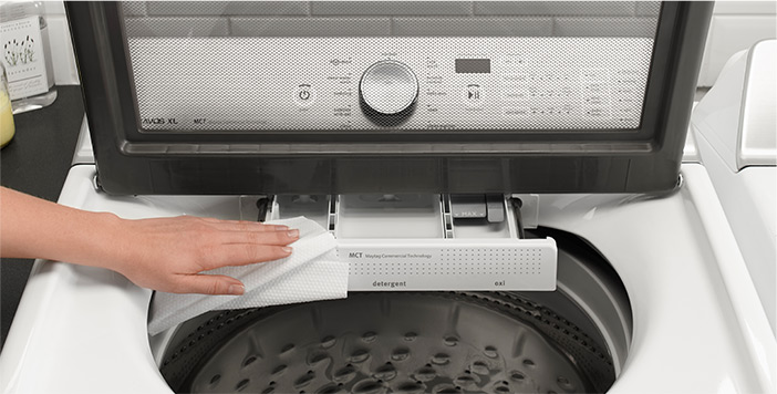 Une main nettoie l'intérieur d'une laveuse à chargement par le haut avec un chiffon. Les commandes de la laveuse sont visibles à travers le hublot de la porte