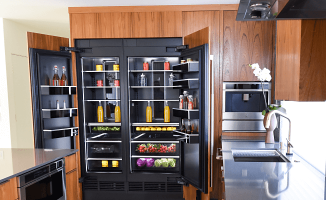 An open JennAir Column Refrigerator filled with fine foods and ingredients in a kitchen with wooden cabinetry. 