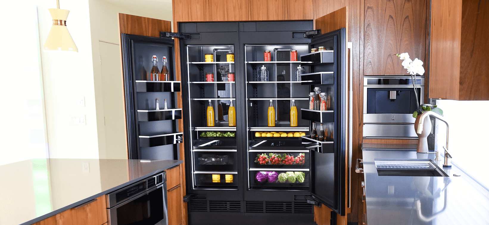 An open JennAir Column Refrigerator filled with fine foods and ingredients in a kitchen with wooden cabinetry. 