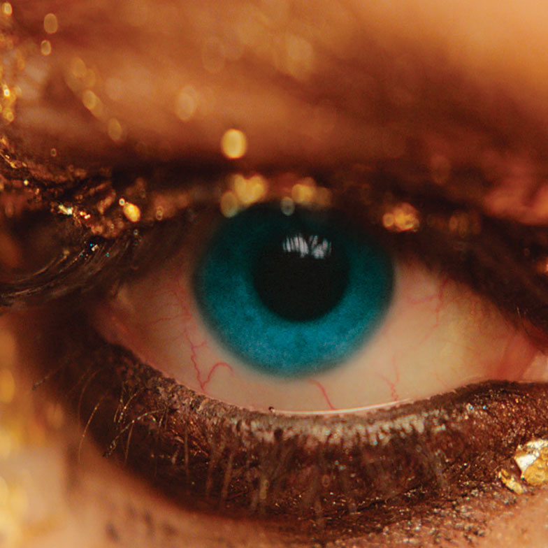 A person with gold makeup and blue eyes looking into the camera. 
