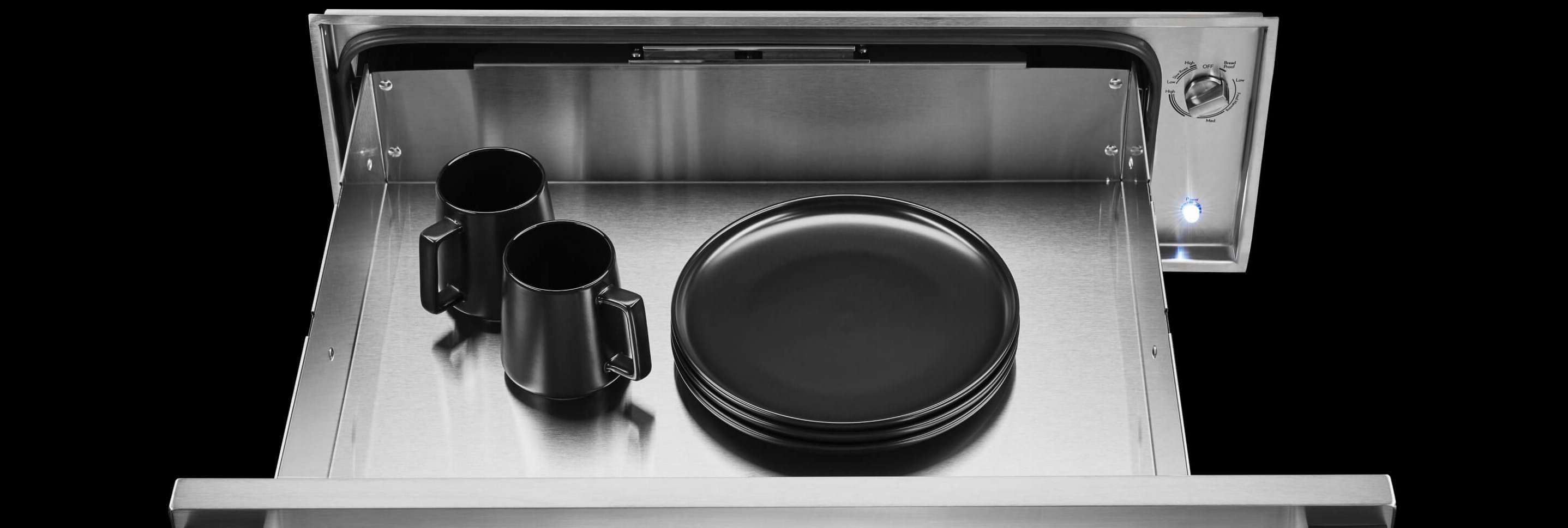 An open JennAir® warming drawer with black plates and mugs inside.