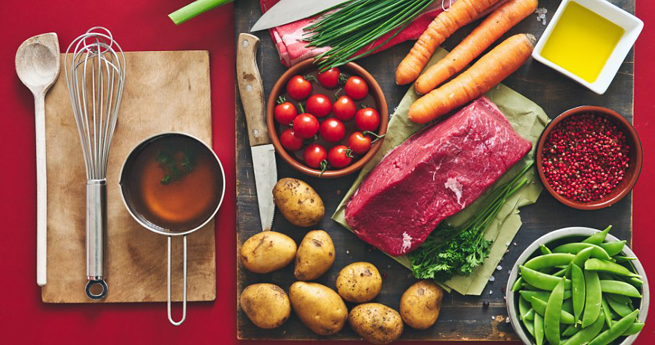 Sur une table rouge se trouvent un linge à vaisselle, une spatule, un fouet et une casserole. À côté se trouve une planche à découper avec un couteau, de la ciboulette, un bol de tomates cerises, des pommes de terre, un morceau de viande, des carottes, un bol de pois mange-tout, un bol d'huile et un bol de pommes grenade