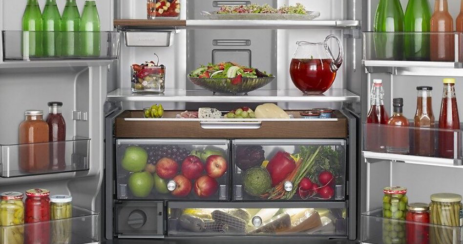 Un réfrigérateur ouvert. Le contenu comprend des bouteilles d'eau, des contenants de jus de fruits, un bol de salade, un pichet de jus de fruits, des bacs à légumes remplis de fruits et légumes tels que des pommes, des poivrons, du céleri et autres, des sauces à salade, des bocaux contenant des olives et des haricots verts et un tiroir contenant du fromage