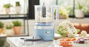 On a counter is a blue KitchenAid food processor with the lid next to it. There is also a cauliflower and a bowl with an orange vegetable. In the background are potted plants on shelves and another potted plant on the windowsill. 