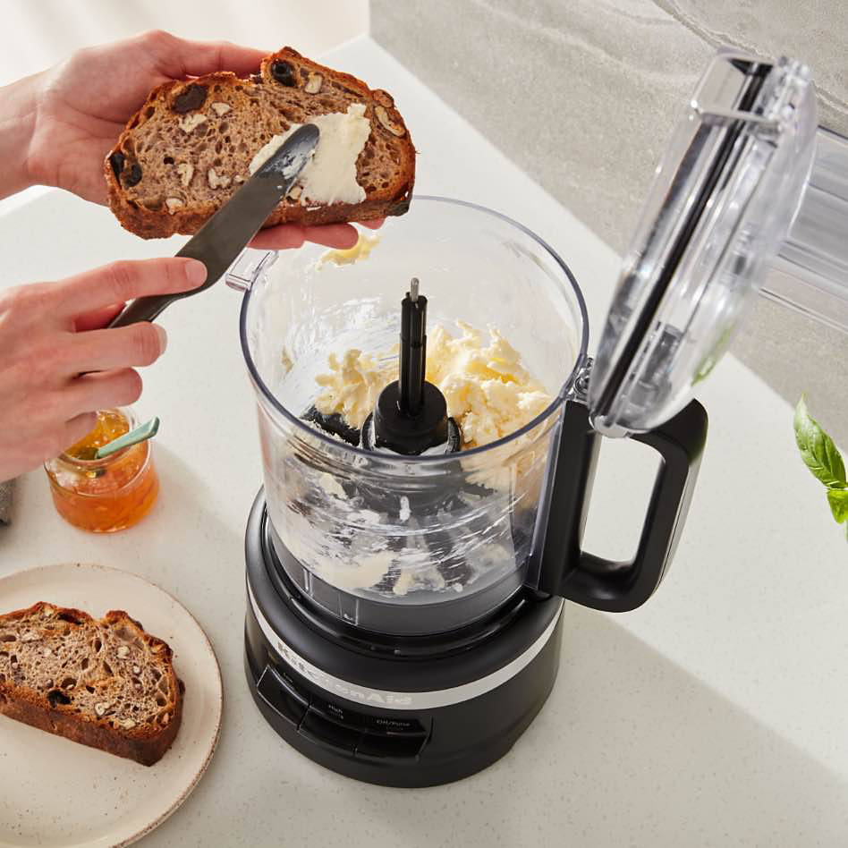 Quelqu'un beurrant un morceau de pain avec du beurre provenant d'un robot culinaire KitchenAid. Une assiette avec une autre tranche de pain se trouve à côté de l'appareil de comptoir, ainsi qu'un petit pot de marmelade