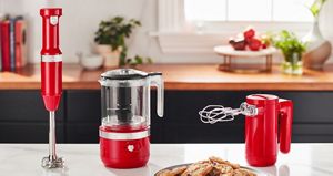 Sur le comptoir, il y a un mélangeur à main KitchenAid, un hachoir KitchenAid et un batteur à main KitchenAid. Devant eux, il y a une assiette de biscuits. Sur le comptoir derrière se trouvent des livres, des plantes en pot, des tomates sur un plateau et des herbes fraîches dans une tasse en verre.