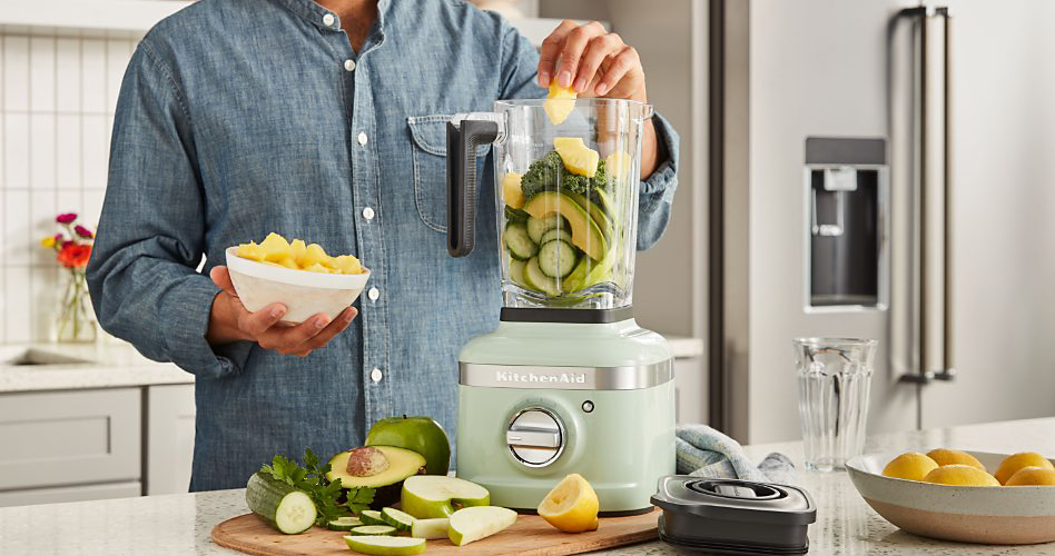 A man adds various vegetables to a mint green KitchenAid blender. In the background: KitchenAid stainless steel French door refrigerator