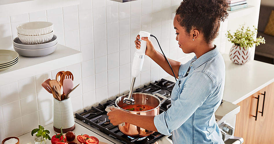 Une femme utilise un mélangeur à main blanc KitchenAid pour mélanger une soupe ou une sauce dans un chaudron sur une cuisinière
