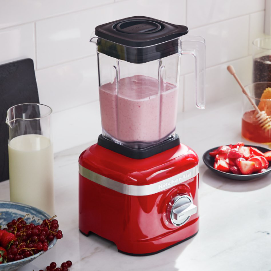 Un mélangeur KitchenAid rouge contenant un smoothie rose sur un comptoir avec divers aliments et articles de cuisine