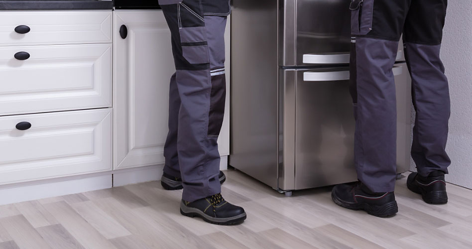Two people in work uniforms in front of a KitchenAid fridge. Behind one of them are white drawers and a white cabinet