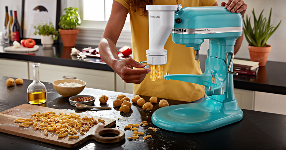Dans une cuisine, une personne vêtue de jaune prépare des pâtes fraîches avec un accessoire pour faire des pâtes sur un batteur sur socle KitchenAid. Sur le comptoir se trouvent une bouteille d'huile transparente, plusieurs boules de pâtes, un bol jaune rempli de farine. Au fond, sur un comptoir, on y voit des bouteilles de vin, des tomates et trois plantes en pot