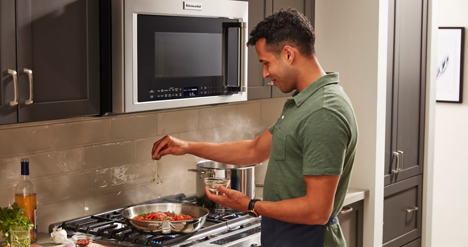 Dans une cuisine, un homme en chemise verte saupoudre des ingrédients dans une casserole qui cuit des ingrédients rouges sur une table de cuisson. Devant lui se trouve un four à micro-ondes avec une bouche d'aération entouré d'armoires grises. Une casserole se trouve également sur la table de cuisson. À côté de la table de cuisson se trouve une bouteille de vin, un bulbe d'ail et des herbes fraîches dans une tasse d'eau