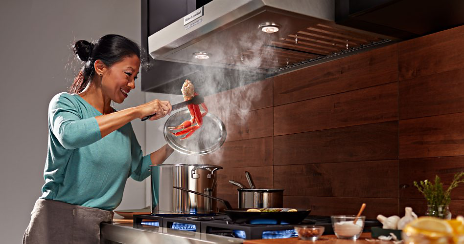 Une femme portant une chemise turquoise utilise des pinces pour ajouter des pattes de crabe dans une casserole fumante sur une table de cuisson. Une casserole plus petite se trouve également sur la table de cuisson. Au-dessus de la table de cuisson, entre des meubles noirs, se trouve une hotte dont les lumières sont allumées. Sur le comptoir à côté de la table de cuisson se trouve un bol d'ail et deux autres bols