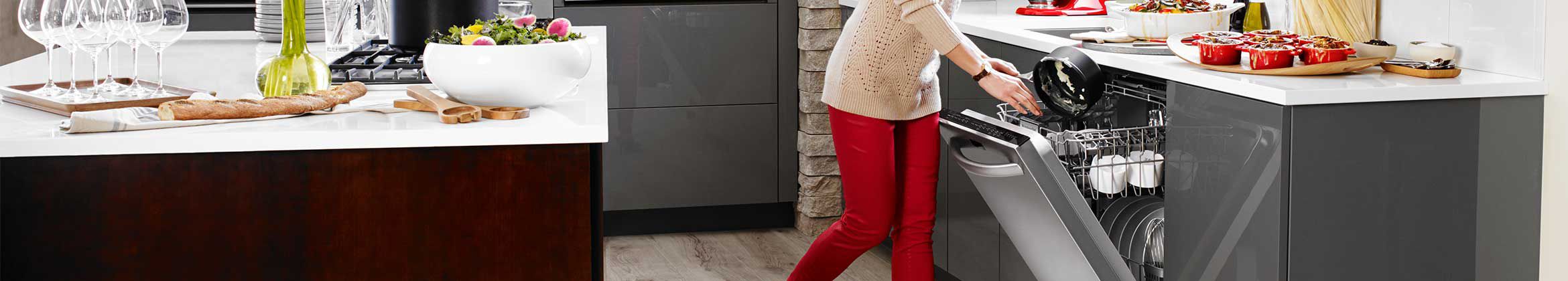 Quelqu'un retire une casserole d'un lave-vaisselle KitchenAid avec la porte ouverte. Sur le comptoir devant se trouvent de petites cocottes et divers autres articles de cuisine. Sur l'îlot derrière se trouvent un saladier rempli de salade, de la nourriture et d'autres articles de cuisine.