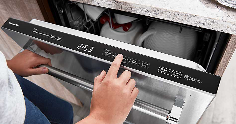 Un lave-vaisselle à commande supérieure KitchenAid avec la porte ouverte. Quelqu'un appuie sur un bouton du panneau de commande. L'heure indique 2 h 53. À l'intérieur du lave-vaisselle se trouvent des tasses et un bol en céramique.