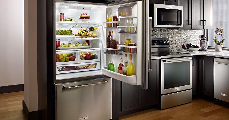 Réfrigérateur à congélateur inférieur KitchenAid avec la porte ouverte. À l'intérieur se trouvent divers fruits et légumes, des bouteilles, des sauces et des boissons. Dans la cuisine se trouvent un four à micro-ondes KitchenAid, un four KitchenAid et un lave-vaisselle KitchenAid.
