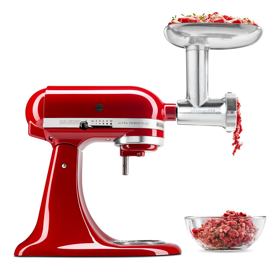 A KitchenAid Stand Mixer with a Metal Food Grinder Attachment. Strands of meat are falling into a bowl that is filled with ground meat. Next to it is a plate of sausage