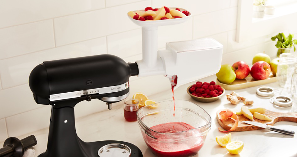 A KitchenAid Stand Mixer with a Fruit and Vegetable Strainer Attachment that is straining fruit into a bowl. On the counter is a plate of apples, a bowl of raspberries, a cutting board with a knife and sliced fruit, cut lemons and fresh herbs