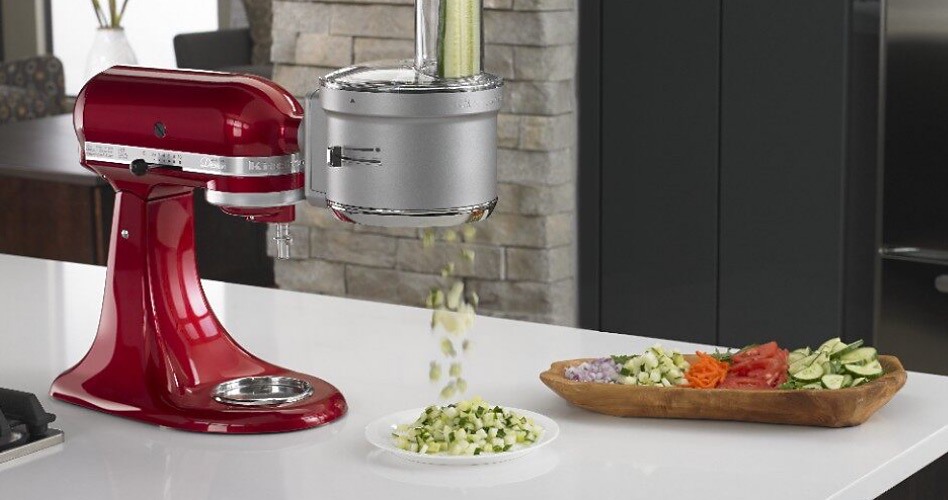 A KitchenAid Stand Mixer with a Food Processor Attachment that is chopping cucumbers onto a plate. There is a tray of sliced tomatoes, onions, carrots and cucumbers on a plate next to it