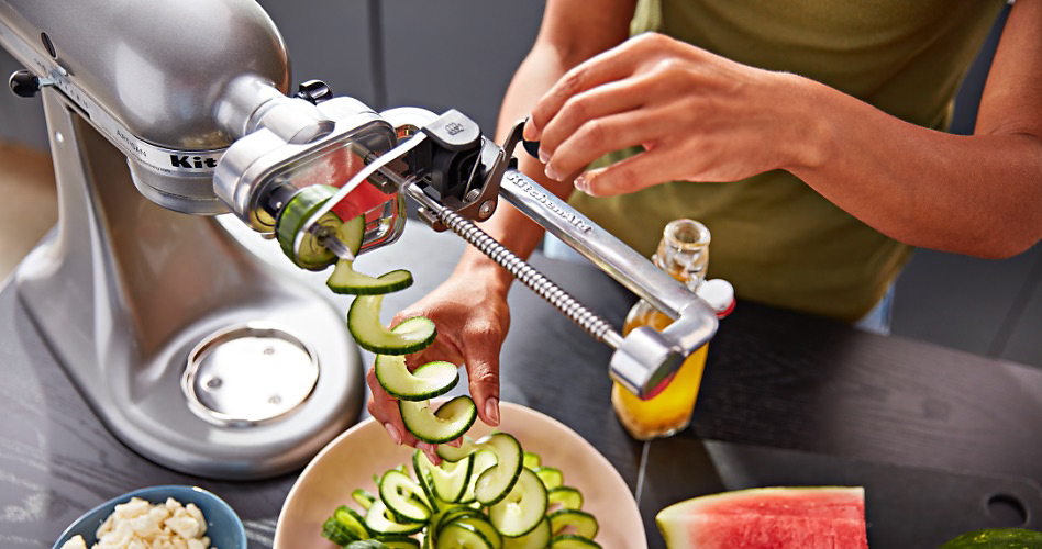 A KitchenAid Stand Mixer with a Spiralizer that is spiralizing cucumbers. On the counter is half a watermelon, a bottle of oil and a bowl of chopped garlic