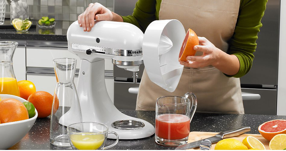 Someone juicing an orange into a glass on a KitchenAid Stand Mixer with a Citrus Juicer attachment. On the counter are a cut grapefruit, oranges, lemons and a bowl of oranges