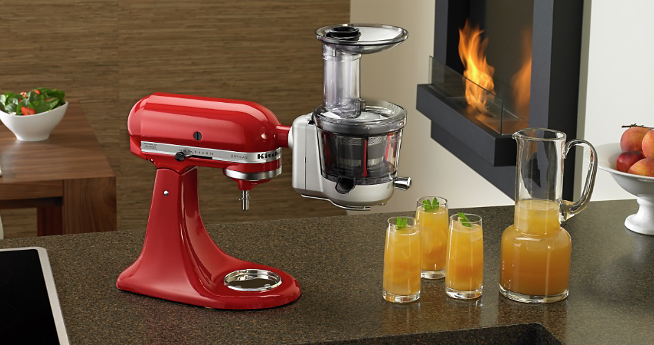 A KitchenAid Stand Mixer with a Slow Juicer & Sauce Attachment. Next to it are three glasses and a pitcher of juice. In the background is a fireplace with a roaring fire
