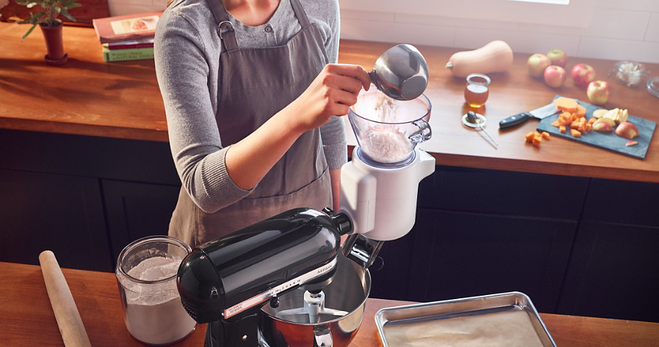 A KitchenAid Stand Mixer with a Sifter + Scale Attachment. Someone is filling the attachment with flour. On the counter is a jar of flour and a rolling pin. In the background are apples, a squash and a cutting board with produce and a knife
