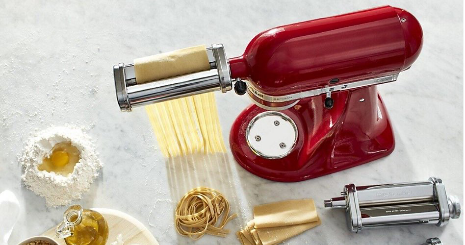 A KitchenAid Stand Mixer with a 3-piece pasta roller and cutter set making pasta strands. Next to it is a flour well with eggs in the centre, a bottle of oil, a nest of pasta, pasta sheets and another pasta roller