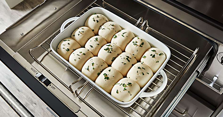Sur une grille dans un tiroir chauffant KitchenAid se trouve un plat de cuisson avec de la pâte crue en forme de rouleaux, garnie d'herbes