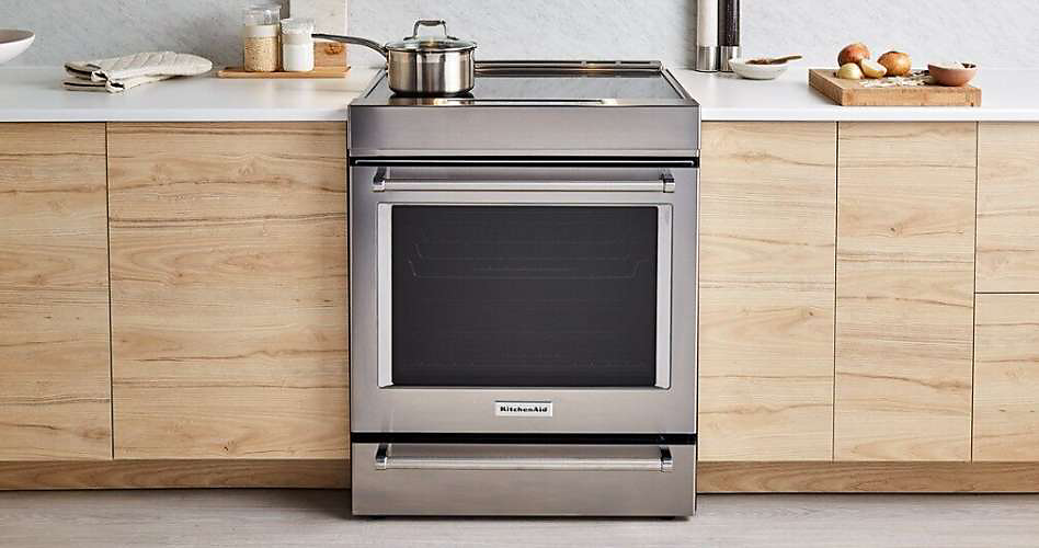 Une cuisinière KitchenAid entourée d'armoires en bois. Sur le comptoir se trouvent un bol à fruits, des gants de cuisine, du sel et une planche à découper avec des fruits et légumes et un petit bol