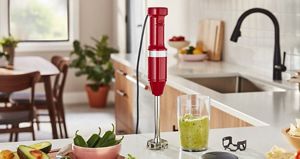 A red KitchenAid immersion hand blender stands upright on a counter. Next to it is a glass filled with guacamole, a bowl of jalapenos and some avocadoes sliced in half.