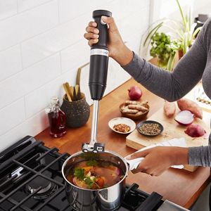 Using a black KitchenAid immersion hand blender, someone purees ingredients as it cooks in a pot on a gas cooktop. Next to them are bowls filled with herbs and spices, another filled with ginger root and a cutting board with sweet potatoes and an onion sliced in half. Behind the cutting board are two potted plants.