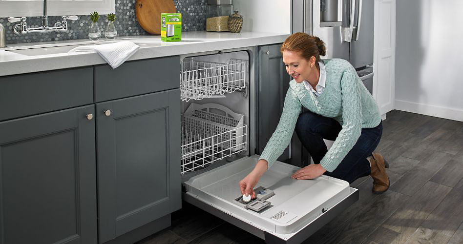 Une femme s'accroupit pour placer une pastille nettoyante affresh dans son lave-vaisselle KitchenAid. Une boîte de tablettes nettoyantes affresh se trouve sur le comptoir. Un torchon est suspendu au-dessus de l'évier. Derrière la femme se trouve un réfrigérateur KitchenAid. 