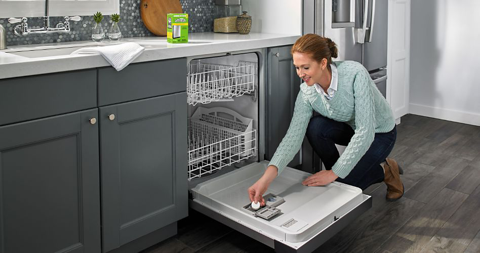 Une femme s'accroupit pour placer une pastille nettoyante affresh dans son lave-vaisselle KitchenAid. Une boîte de tablettes nettoyantes affresh se trouve sur le comptoir. Un torchon est suspendu au-dessus de l'évier. Derrière la femme se trouve un réfrigérateur KitchenAid.
