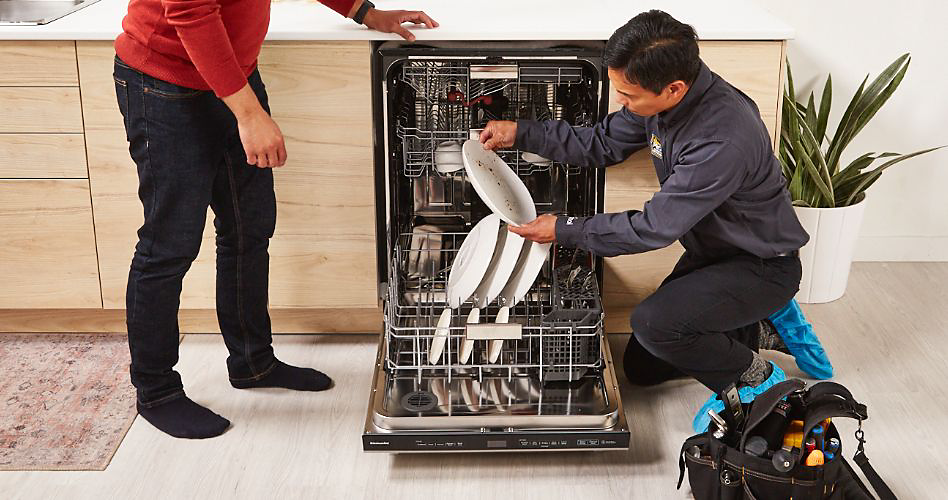 Un réparateur inspecte une assiette qui sort d'un lave-vaisselle KitchenAid. Ses outils sont à côté de lui et il y a une plante derrière lui. Un autre homme en jeans et chemise rouge se trouve de l'autre côté du lave-vaisselle. 