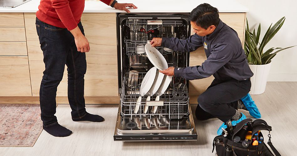 Un réparateur inspecte une assiette qui sort d'un lave-vaisselle KitchenAid. Ses outils sont à côté de lui et il y a une plante derrière lui. Un autre homme en jeans et chemise rouge se trouve de l'autre côté du lave-vaisselle.