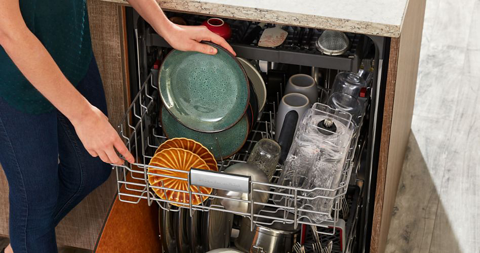 Quelqu'un place une assiette sur le panier d'un lave-vaisselle. On y trouve également des assiettes, des tasses, des verres et d'autres objets.