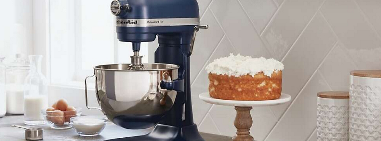 Un batteur sur socle KitchenAid bleu foncé est posé sur un comptoir. Sur le comptoir se trouvent également un gâteau avec un glaçage blanc sur un plateau à gâteau, un bol d'œufs, un bol contenant un ingrédient blanc, deux récipients blancs et une bouteille de lait.