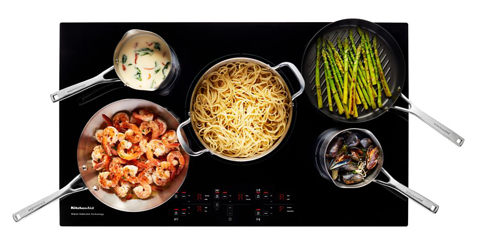 Divers aliments cuisant sur une cuisinière en vitrocéramique, tels que des crevettes, des asperges, des pâtes, et plus.