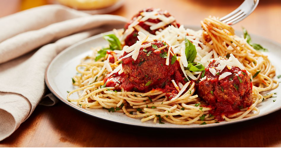 Une fourchette sort les pâtes d'un bol de spaghetti et de boulettes de viande garni de persil et de parmesan.  Une serviette de table en tissu se trouve à côté de l'assiette