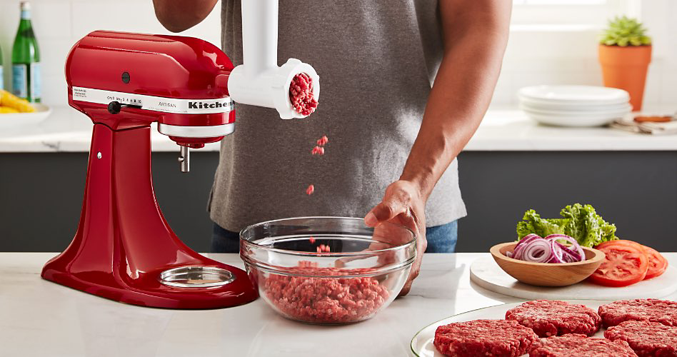 Une personne broie de la viande dans un bol en verre à l'aide d'un hachoir à viande rouge. Sur le comptoir se trouvent une assiette de galettes de hamburger et un bol d'oignons rouges, de tomates et de laitue. Sur un comptoir en arrière-plan se trouvent une plante en pot, une bouteille d'eau gazeuse et une pile d'assiettes