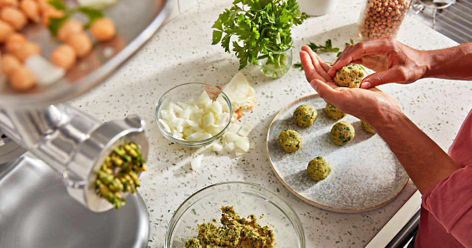 Sur un comptoir, quelqu'un roule de la viande en boulettes. Sur le comptoir se trouve également un hachoir à viande d'où sort de la viande, un bol en verre et un récipient en verre contenant des herbes fraîches