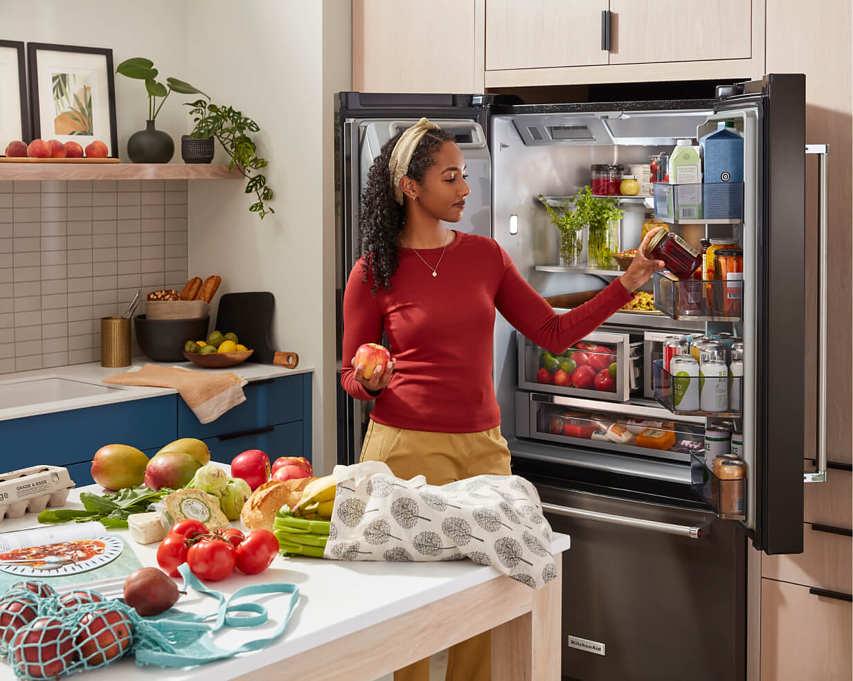An open KitchenAid® refrigerator with a person placing free produce and fruits into the different shelves and drawers. 
