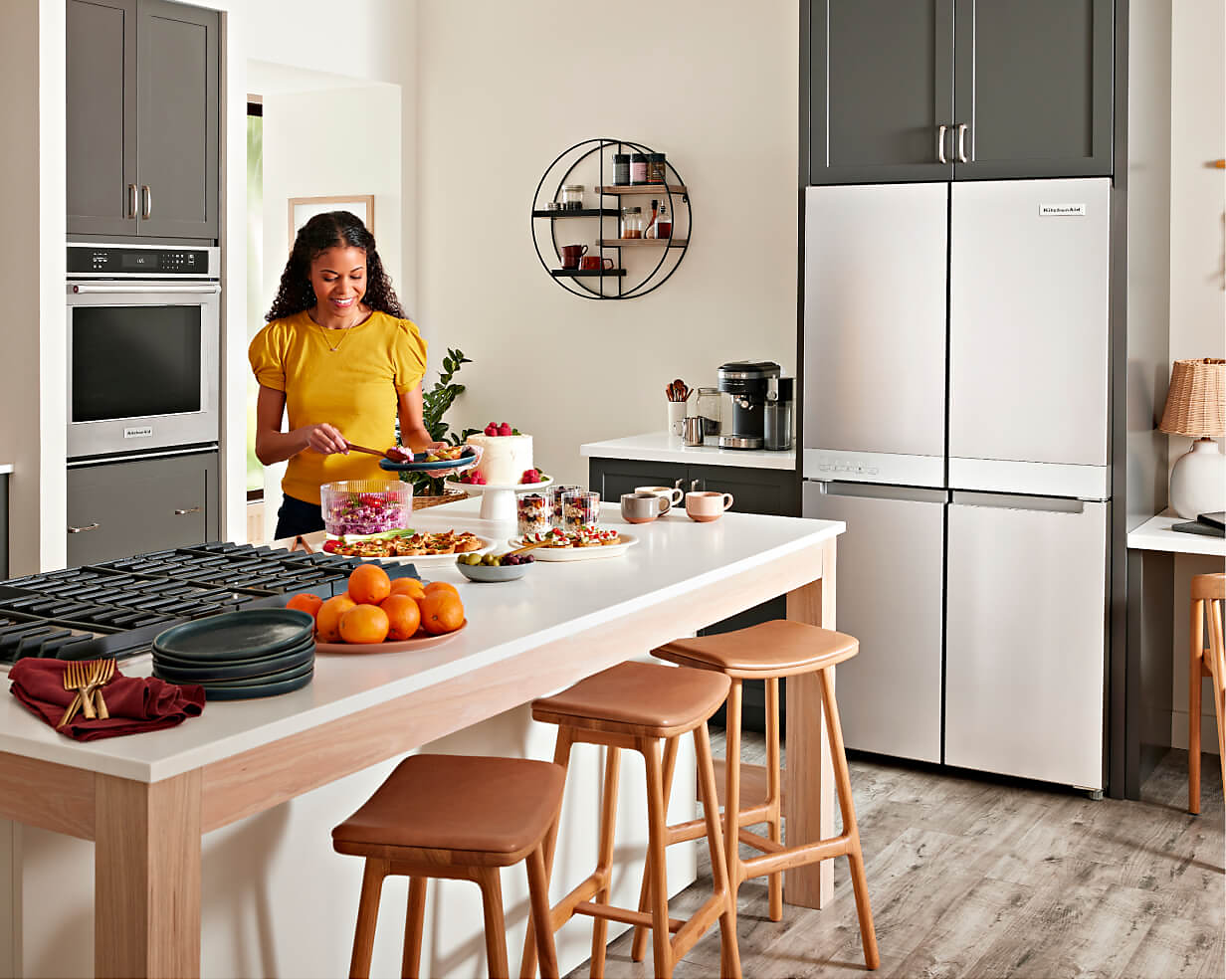 A KitchenAid® four-door freestanding refrigerator in a stylish kitchen filled with a suite of KitchenAid® appliances and a person preparing food for a gathering.