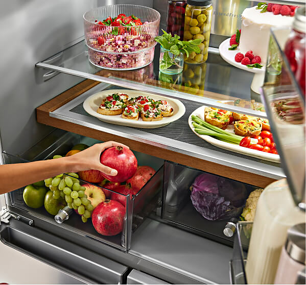 The interior of a refrigerator with different prepared appetizers on party platters and a person placing fresh fruit into a crisper drawer.