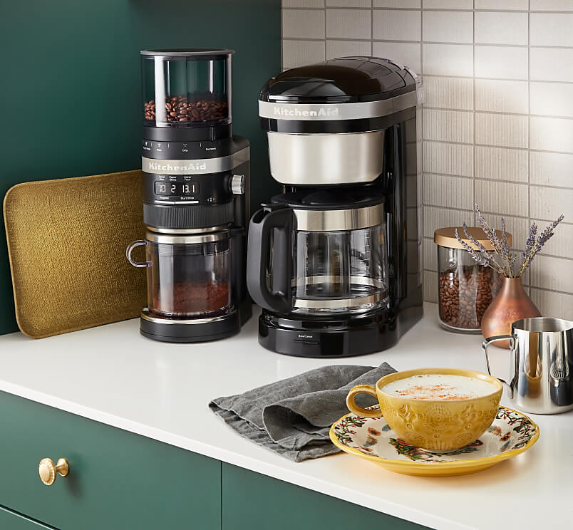 A KitchenAid® grinder next to a KitchenAid® coffee maker on a counter with a cup of coffee.