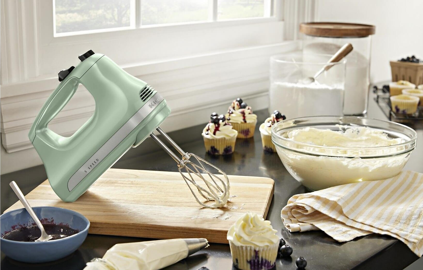 A KitchenAid® Hand Mixer on a countertop with a bowl of frosting and cupcakes.