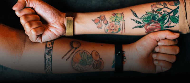 Two arms side by side, showing their gardening and food tattoos.